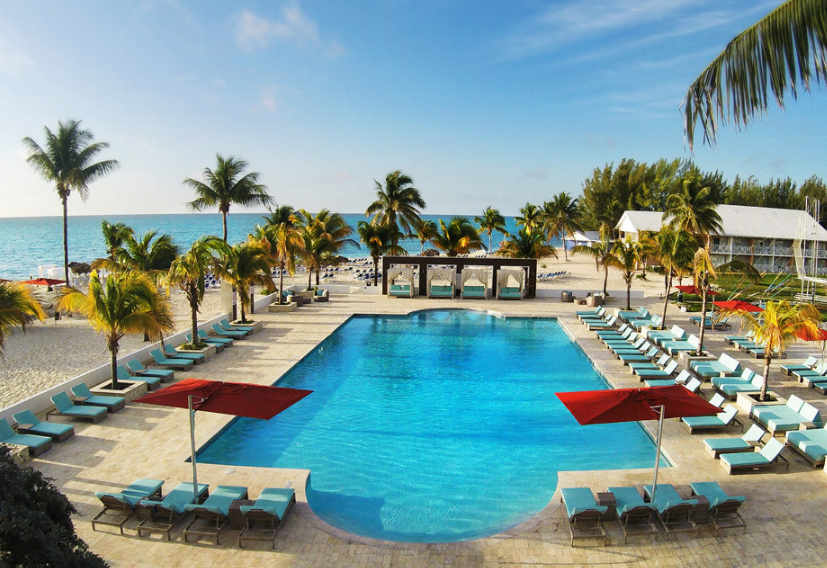 A sunny tropical resort features a large swimming pool, beach loungers, palm trees, and the sea in the background.