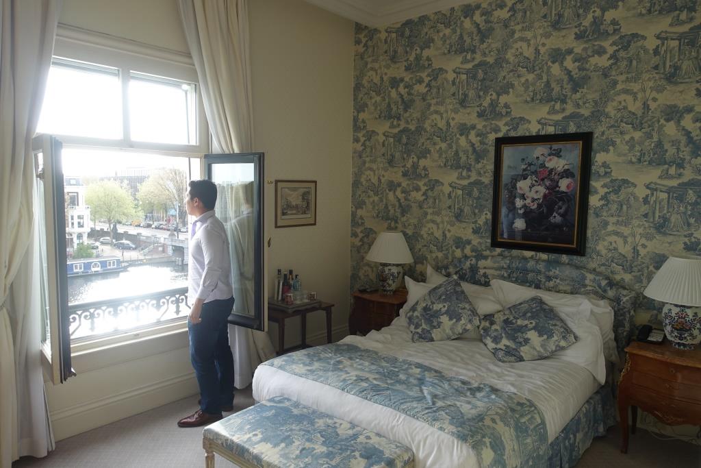 A man looks out an open window in a hotel room with blue and white toile decor, overlooking a city canal.