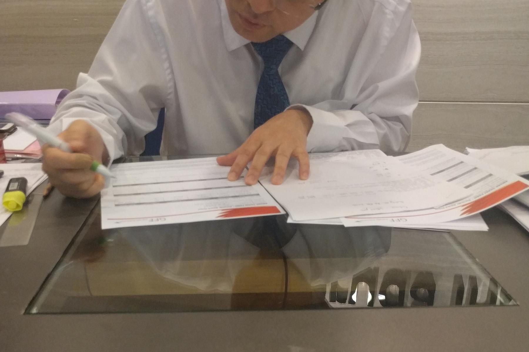 A person in a white shirt and tie fills out documents at a desk.