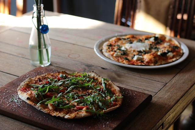 Two pizzas, one topped with arugula and the other with cheese and spinach, sit on a rustic wooden table.