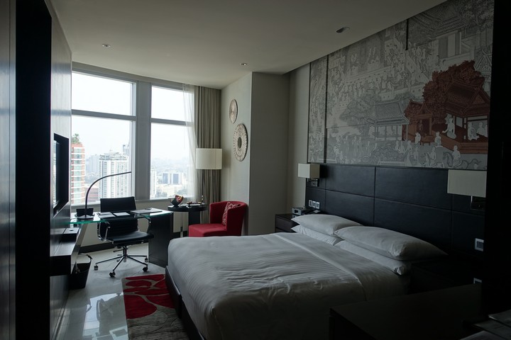 A luxurious hotel room with a king-sized bed, a desk by a window overlooking a city, and a traditional mural above the bed.