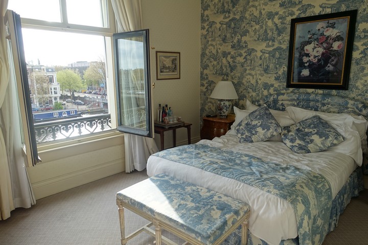 A hotel room with blue and white toile wallpaper and bedding, and an open window revealing a canal view.
